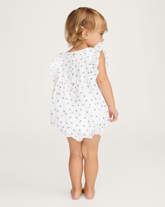 A toddler with blonde hair stands barefoot, facing away, wearing the Baby’s Twill Ruffled Romper in Anchors Away White—a sleeveless romper featuring small colorful anchor prints and ruffled shoulders—set against a plain light background.