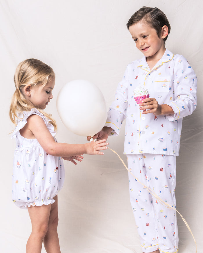Two kids, dressed in matching Petite Plumes yarn-dyed cotton pajamas, are together. The girl holds a white balloon, while the boy has a sprinkle-topped cupcake. A neutral backdrop sets the scene for this cozy sleepwear moment featuring Babys Twill Ruffled Romper in Birthday Wishes.