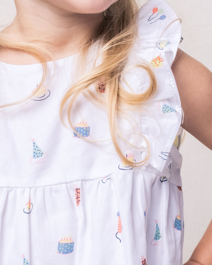 A young girl in a Petite Plume Babys Twill Ruffled Romper, crafted from soft yarn-dyed cotton with party-themed prints of cakes, balloons, and hats. Her long blond curls fall gently over the rompers ruffled sleeves, highlighting her outfits playful elegance.