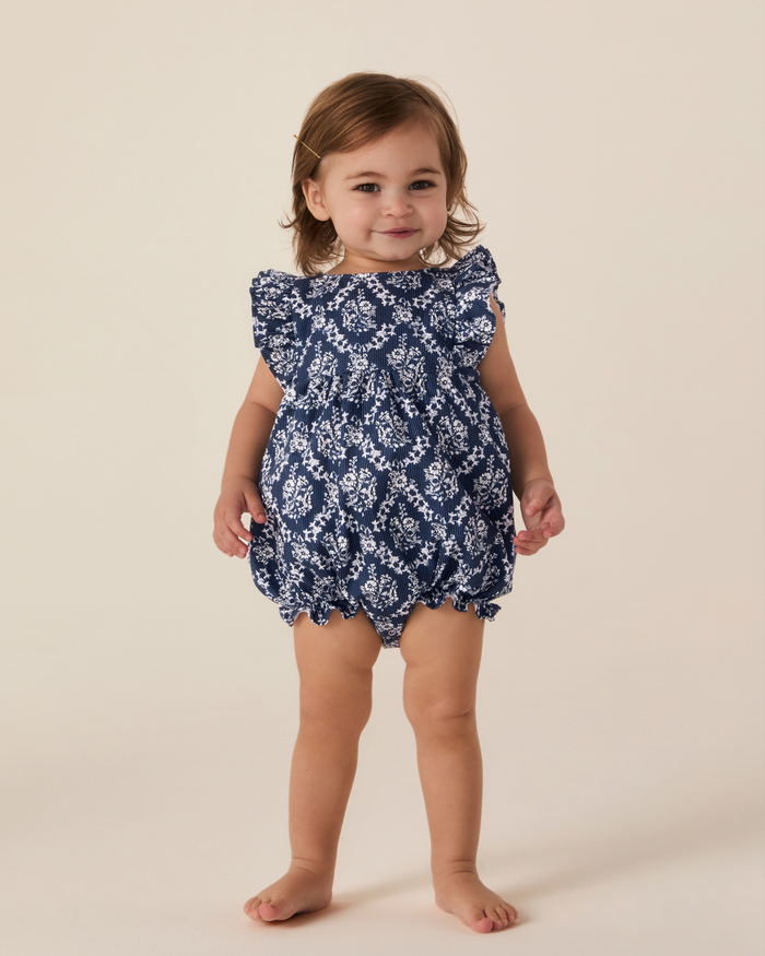 A young toddler with brown hair stands barefoot, smiling at the camera in the Baby’s Twill Ruffled Romper in Innkeeper Trellis against a plain light background.