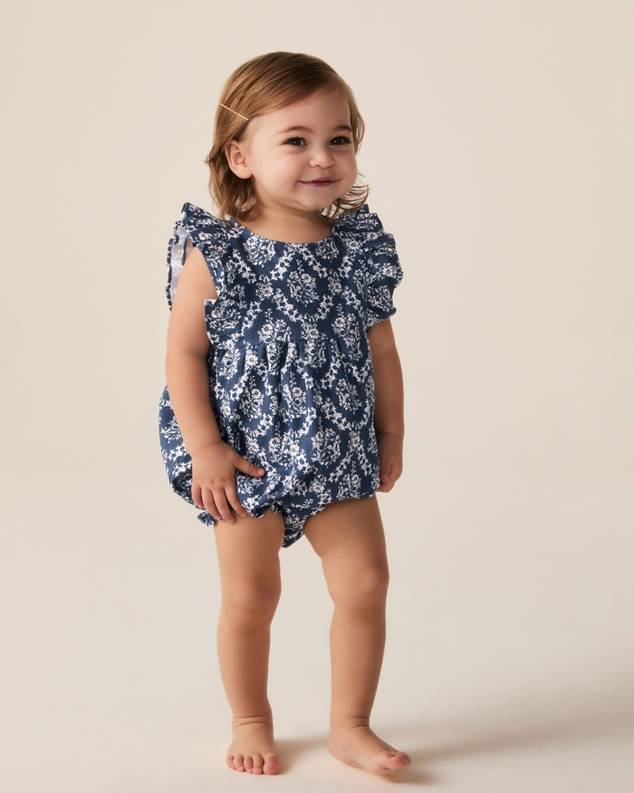 A toddler with light brown hair wears the Baby’s Twill Ruffled Romper in Innkeeper Trellis, featuring blue and white patterns and ruffled sleeves, standing barefoot on a light-colored surface while smiling at the camera.