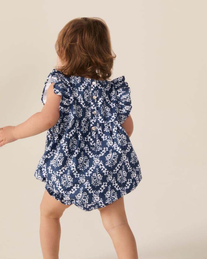 A toddler wearing the Baby’s Twill Ruffled Romper in Innkeeper Trellis walks away from the camera against a plain, light-colored background.