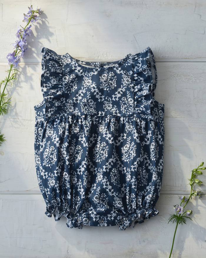 Baby’s Twill Ruffled Romper in Innkeeper Trellis, featuring a blue and white pattern with ruffled sleeves, displayed flat on a light wooden surface, accented by purple and green flowers positioned at the top left and bottom right corners.