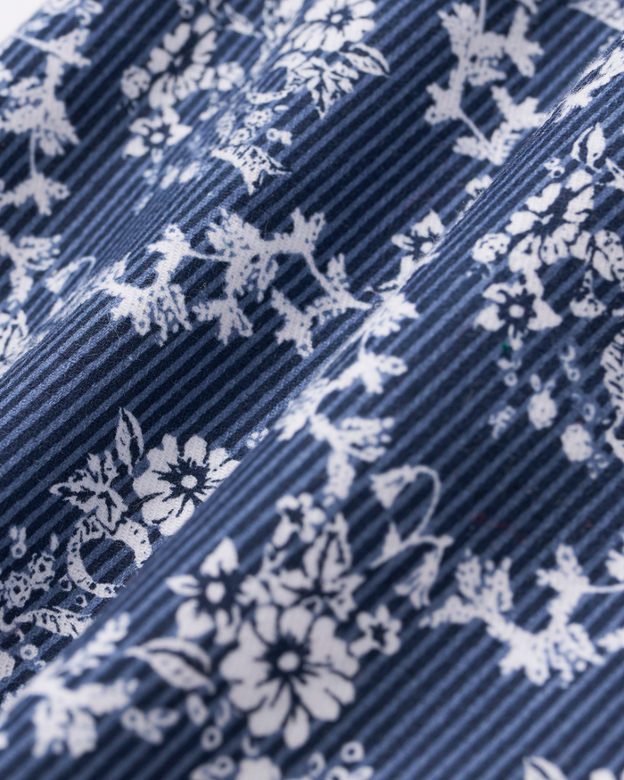 Close-up of the Baby’s Twill Ruffled Romper in Innkeeper Trellis, featuring blue fabric with thin white stripes and a delicate white floral pattern, highlighting soft folds and intricate design details.