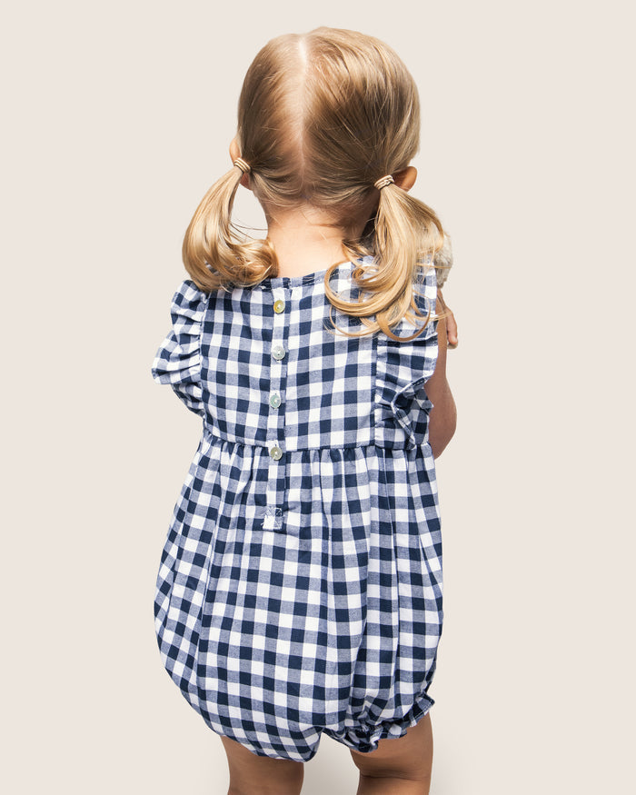 A toddler with pigtails, dressed in Petite Plumes Babys Twill Ruffled Romper in Navy Gingham, faces away from the camera. This yarn-dyed cotton romper features back buttons and ruffled sleeves for comfort and style.