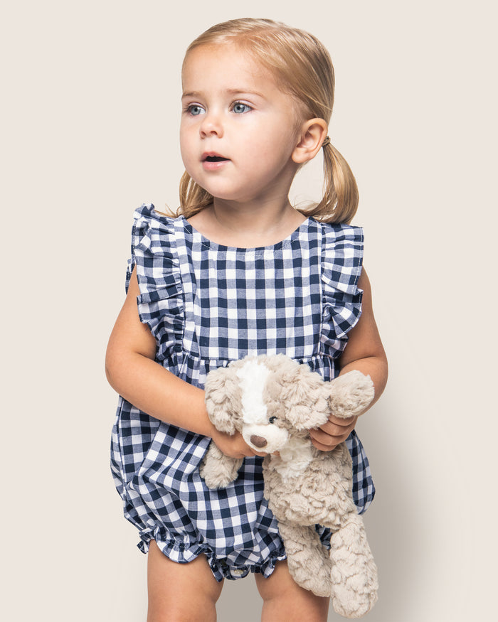 A young blonde child with pigtails wears Petite Plumes Babys Twill Ruffled Romper in Navy Gingham, ideal for stylish sleepwear. The child holds a beige stuffed toy dog and gazes slightly to the side against a light background.