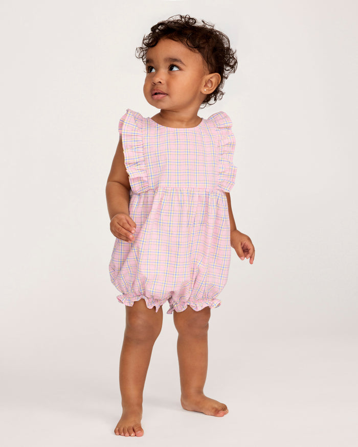 A toddler with curly hair wears the Baby's Twill Ruffled Romper in Peony Plaid, featuring ruffled sleeves, standing barefoot against a plain light background and looking slightly to the side.