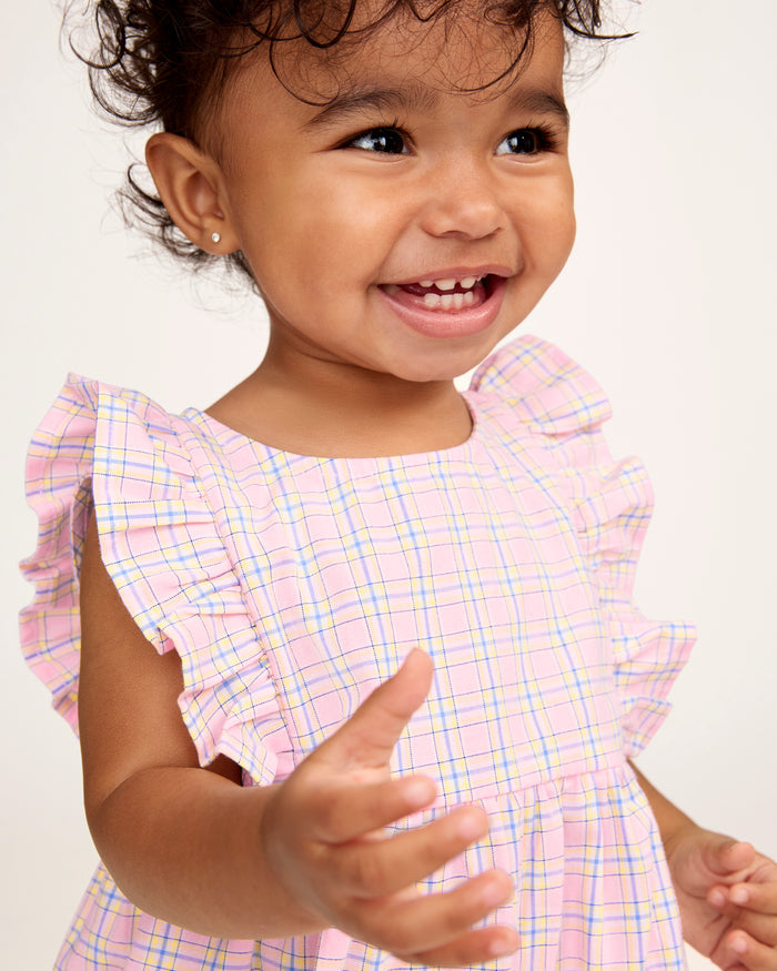 A toddler with curly hair wears the Baby's Twill Ruffled Romper in Peony Plaid, featuring ruffled sleeves, standing barefoot against a plain light background and looking slightly to the side.