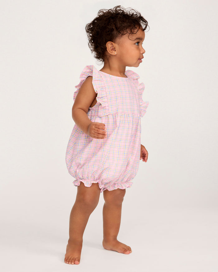 A toddler with curly hair wears the Baby's Twill Ruffled Romper in Peony Plaid, featuring ruffled sleeves, standing barefoot against a plain light background and looking slightly to the side.