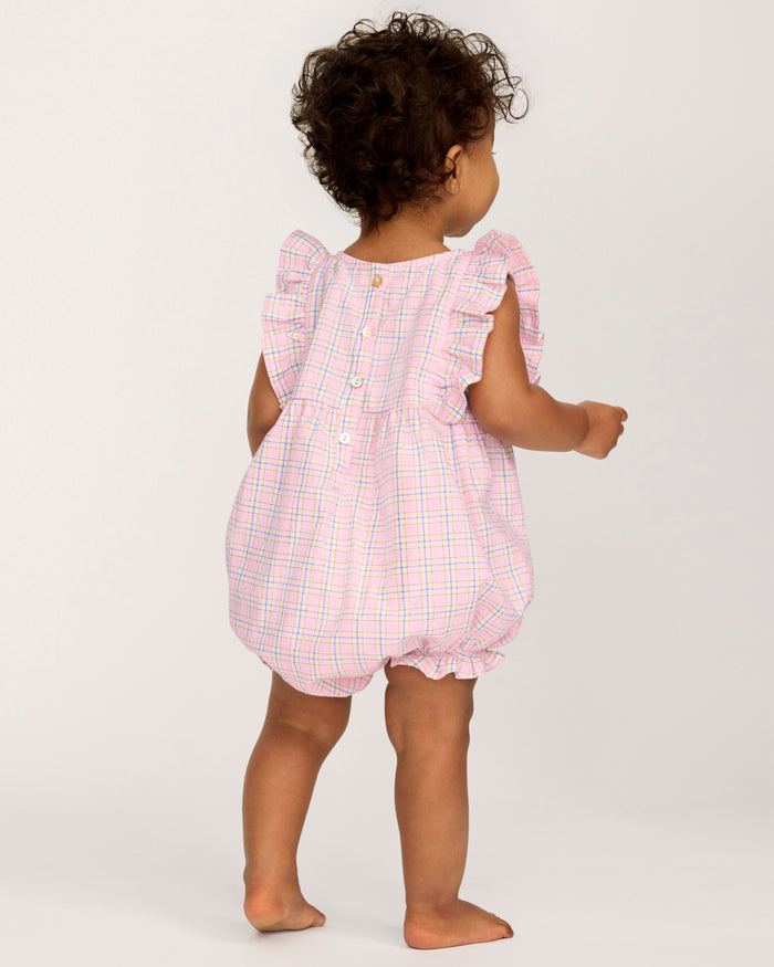 A toddler with curly hair wears the Baby's Twill Ruffled Romper in Peony Plaid, featuring ruffled sleeves, standing barefoot against a plain light background and looking slightly to the side.