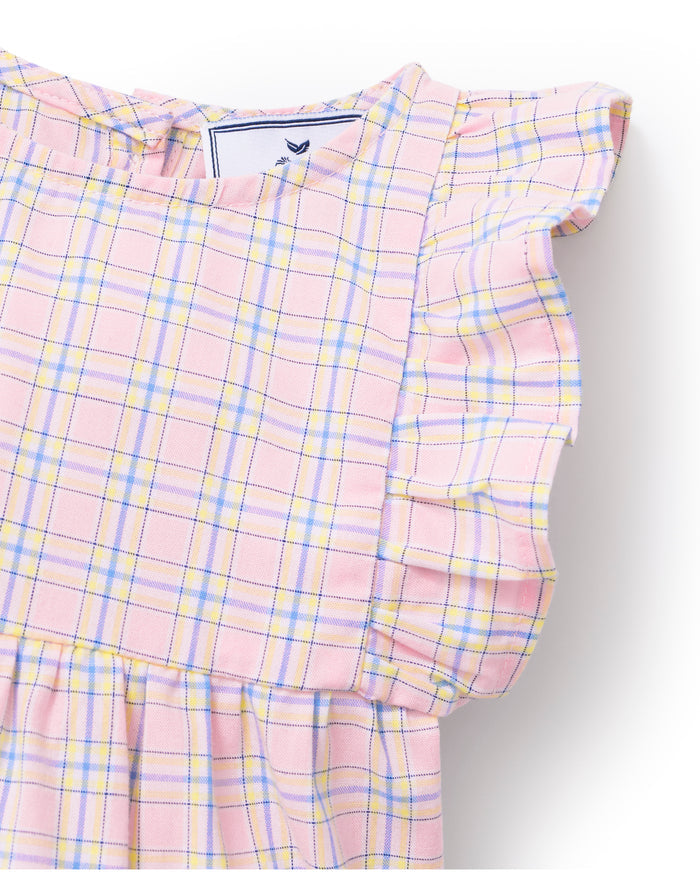 A toddler with curly hair wears the Baby's Twill Ruffled Romper in Peony Plaid, featuring ruffled sleeves, standing barefoot against a plain light background and looking slightly to the side.