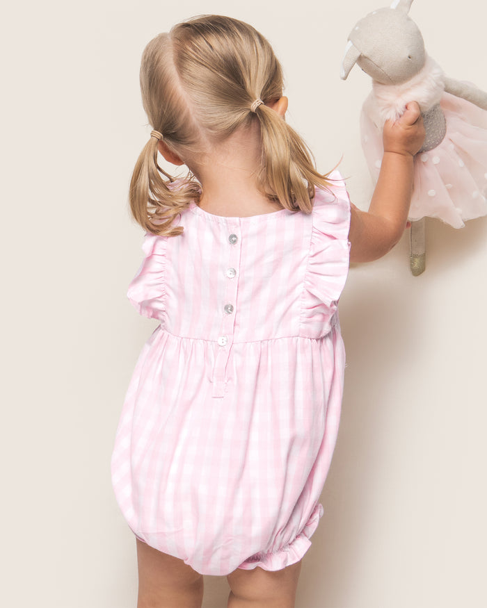 A toddler with light brown pigtails faces away, holding a gray stuffed animal in a pink skirt. She wears the Petite Plume Babys Twill Ruffled Romper in Pink Gingham—a cozy sleepwear piece—buttoned down the back.