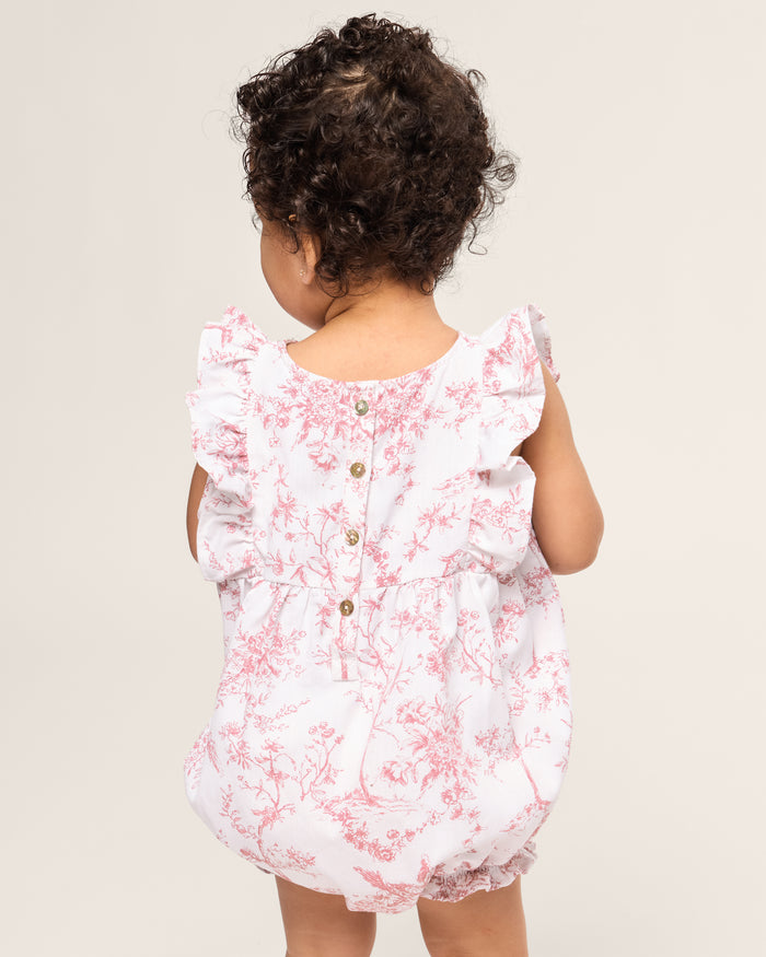 A toddler with curly hair wears Petite Plumes Babys Twill Ruffled Romper in Pink Timeless Toile, featuring floral patterns and ruffled sleeves. The romper has buttons down the back, and the child stands facing away against a plain background.