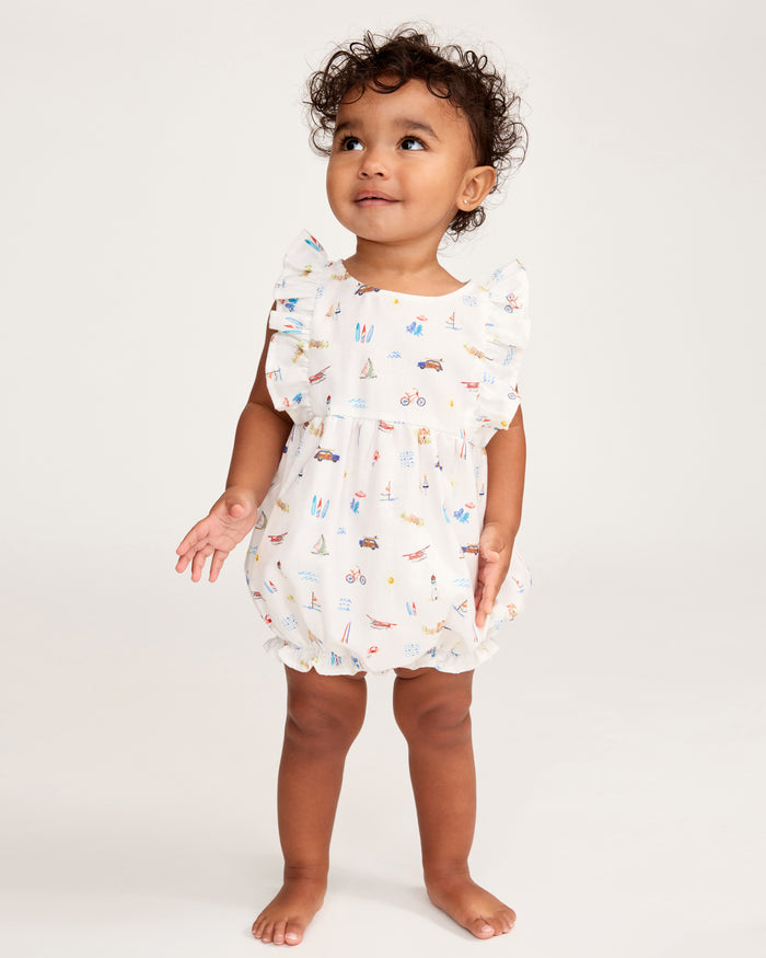 A toddler with curly hair stands barefoot on a light background, smiling and looking up with arms slightly out to the sides, dressed in the Baby’s Twill Ruffled Romper in Summer Nostalgia.