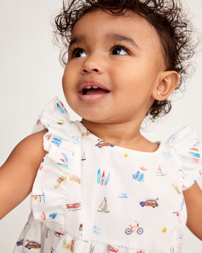 A toddler with curly hair, wearing the Baby’s Twill Ruffled Romper in Summer Nostalgia—featuring a white fabric adorned with colorful beach-themed prints and ruffled sleeves—smiles slightly as she looks up, small gold earrings glinting against a plain background.