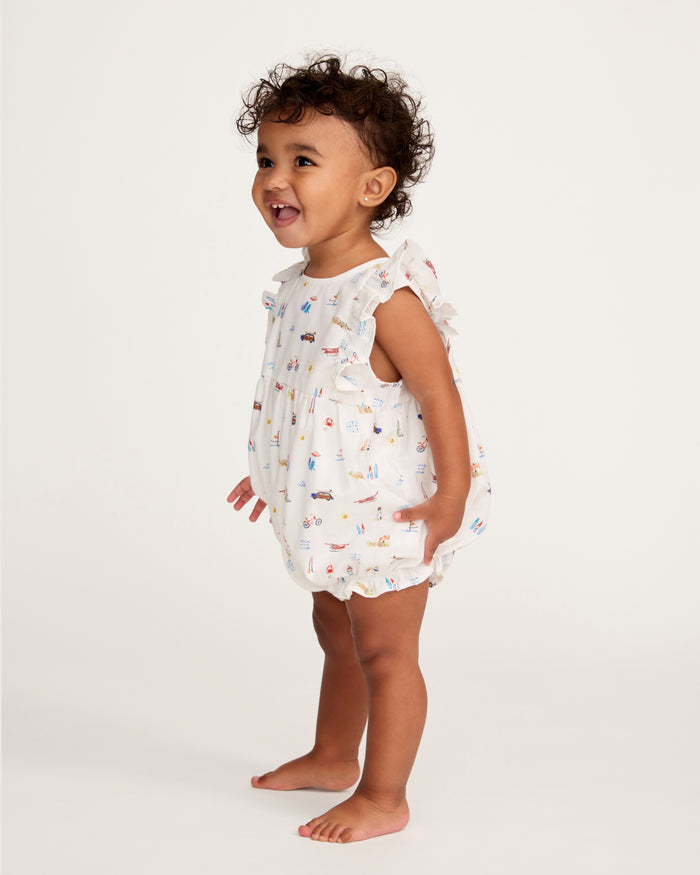 A smiling toddler with curly hair stands barefoot against a plain light background, wearing the Baby’s Twill Ruffled Romper in Summer Nostalgia, adorned with colorful illustrations.