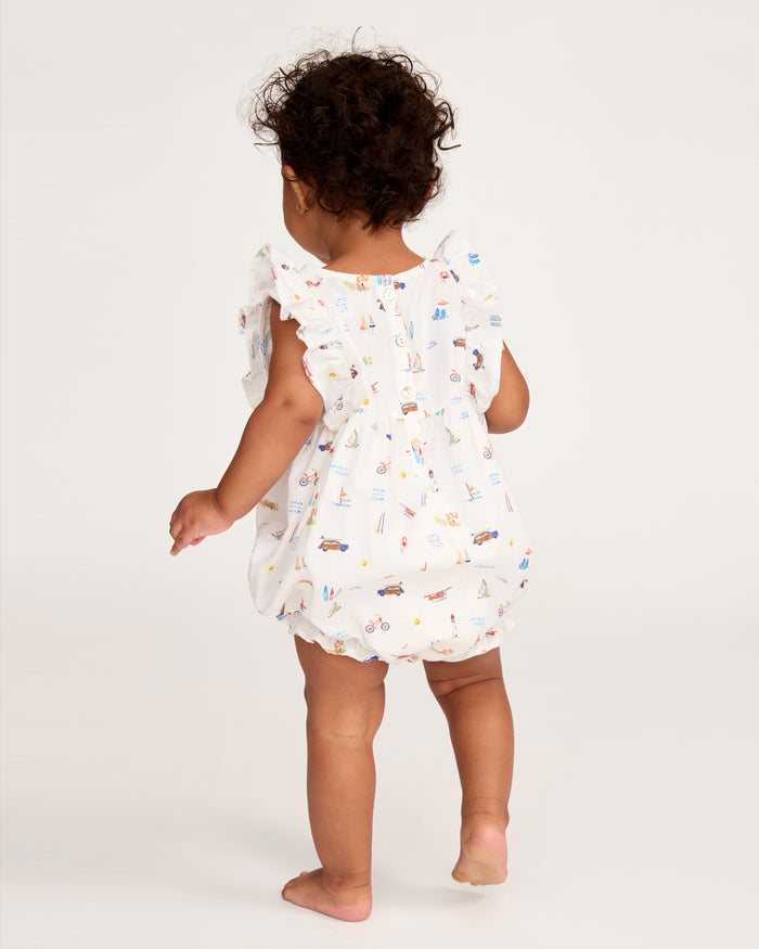 A toddler with curly hair stands barefoot, facing away on a plain light background, wearing the Baby’s Twill Ruffled Romper in Summer Nostalgia—a white romper adorned with colorful illustrations and ruffled details.