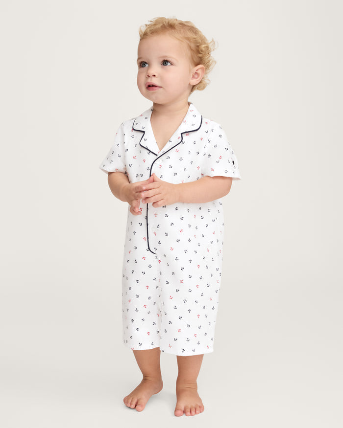 A toddler with curly blond hair stands barefoot, looking slightly upward, dressed in the Baby’s Twill Summer Romper in Anchors Away White—a white short-sleeve romper featuring a small anchor print and navy piping.