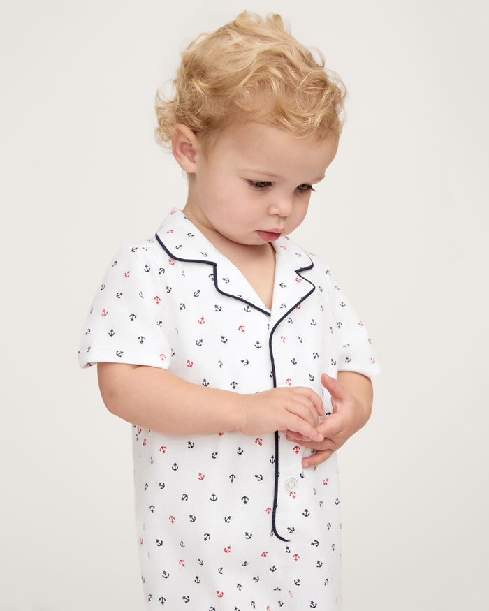 A toddler with curly blond hair wears the Baby’s Twill Summer Romper in Anchors Away White, featuring a small anchor print and dark piping, looking down while touching their fingers. The background is plain and light-colored.