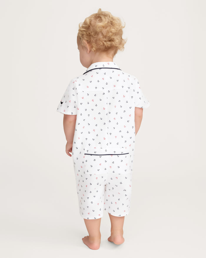 A toddler with curly blond hair stands barefoot, facing away, wearing the Baby’s Twill Summer Romper in Anchors Away White, featuring a small anchor pattern and navy blue piping, on a plain light background.