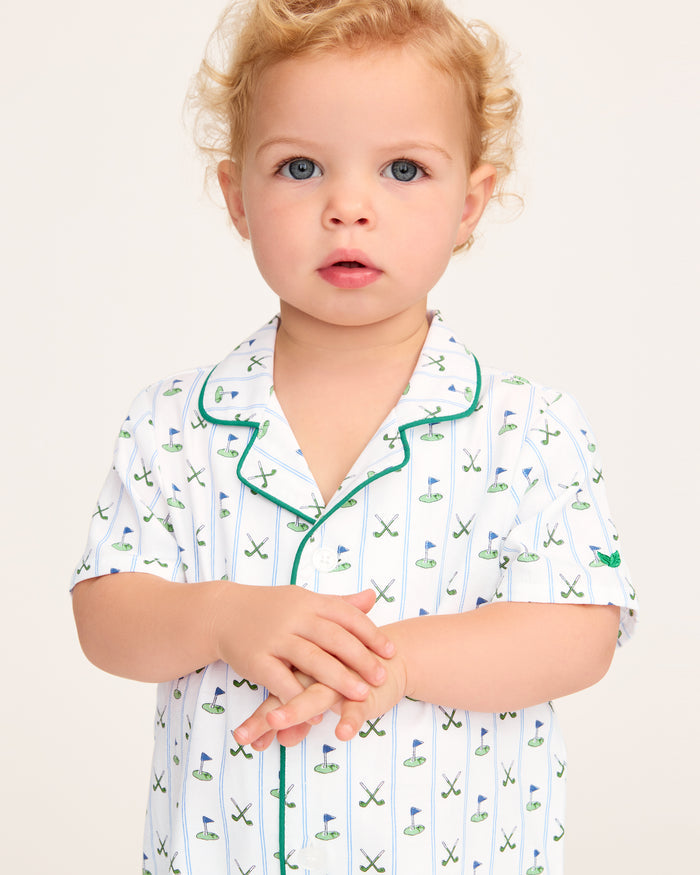 The Baby's Twill Summer Romper in Fairway Stripe is a short-sleeved, button-down romper with green piping, decorated with golf clubs and holes on white fabric with blue vertical stripes.