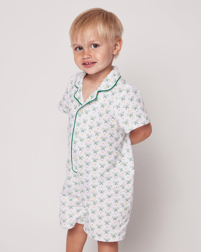 A young child with blonde hair wears the luxurious cotton Babys Twill Summer Romper in Match Point by Petite Plume, featuring green tennis racket patterns and green piping. The child is smiling with hands behind their back against a plain backdrop.