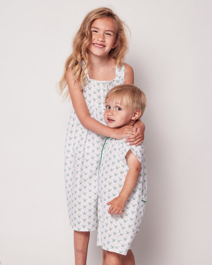 Two young children smile at the camera against a plain background. The older one with long hair hugs the younger one with short hair, both dressed in matching Petite Plume sleepwear adorned in a subtle green floral pattern reminiscent of Babys Twill Summer Romper in Match Point.