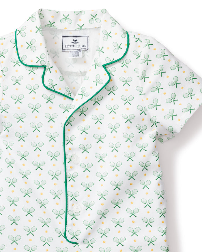 The Babys Twill Summer Romper in Match Point by Petite Plume exudes a classic sport sleepwear vibe with its green tennis racket and yellow ball print. It features green piping on the collar and button line, crafted from luxurious cotton twill with an inner collar label for added detail.