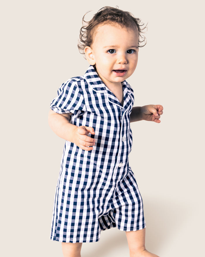 A young child wearing Petite Plumes Babys Twill Summer Romper in Navy Gingham smiles, mid-step, against a light background with curly, tousled hair and one arm bent while the other is slightly extended.