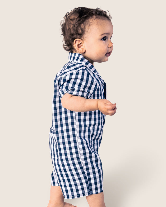 A toddler in Petite Plumes Babys Twill Summer Romper in Navy Gingham stands against a plain background. The childs curly hair and side profile are visible, highlighting the classic charm of this stylish piece.