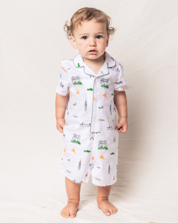 A young child in a Petite Plume Babys Twill Summer Romper, featuring colorful New York landmarks, stands barefoot against a light background gazing slightly upwards with a neutral expression. The romper is woven from soft yarn-dyed cotton and offers flame retardancy.