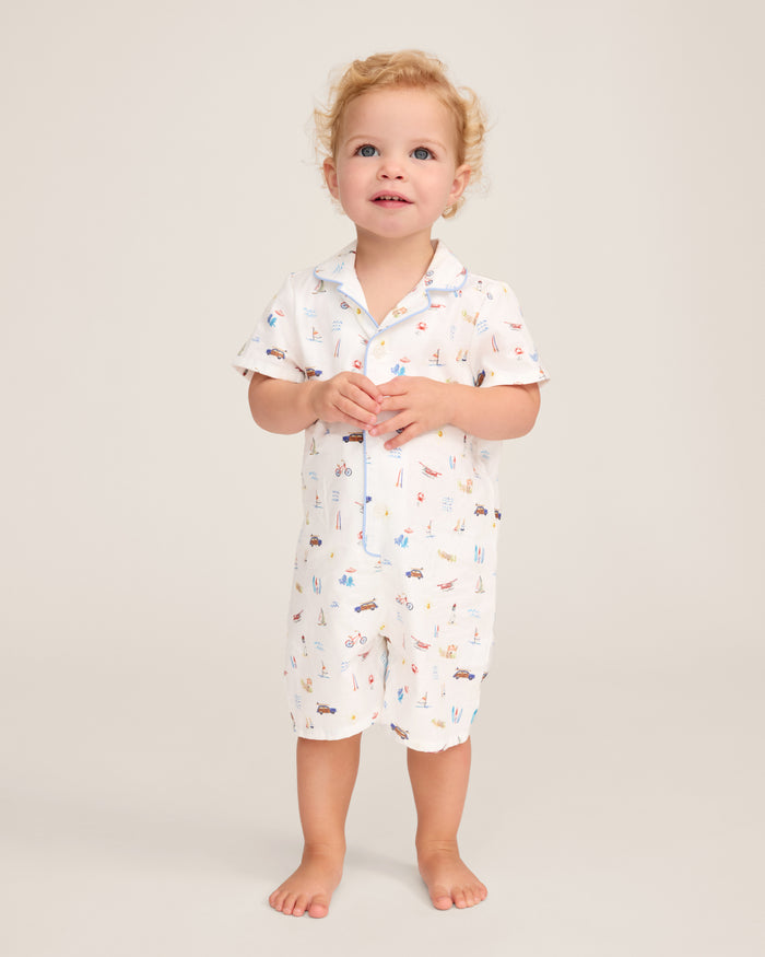 A young child with curly blonde hair stands barefoot against a plain light background, wearing the Baby’s Twill Summer Romper in Summer Nostalgia—a white short-sleeve romper adorned with colorful beach-themed illustrations.