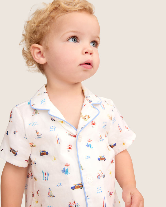A toddler with curly blond hair wears the Baby’s Twill Summer Romper in Summer Nostalgia—featuring a white background adorned with vibrant illustrations of cars, bicycles, surfboards, and sailboats—while looking slightly upward against a plain backdrop.