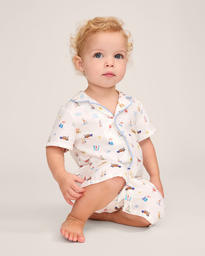 A young child with curly blonde hair sits barefoot on the floor, wearing the Baby’s Twill Summer Romper in Summer Nostalgia—white with colorful illustrations of cars and surfboards—against a plain light background.