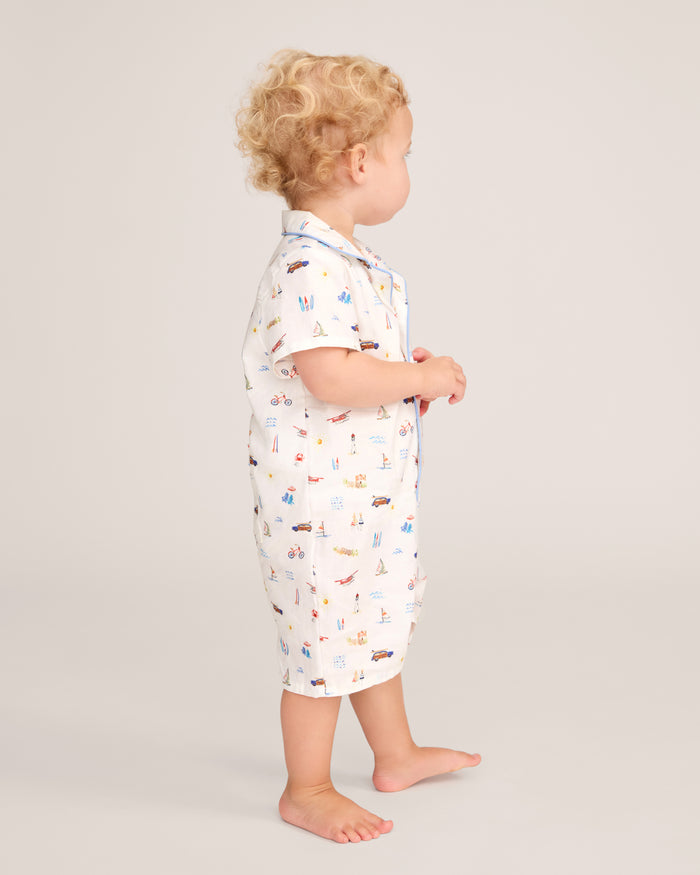A young child with curly blonde hair stands barefoot on a plain light background, facing sideways, wearing the Baby’s Twill Summer Romper in Summer Nostalgia with its colorful prints.