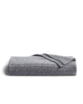 The Petite Plume Cashmere Cable Knit Throw in Dark Grey, elegantly textured with a ribbed edge, is displayed against white. This cozy, giftable piece enhances any home decor with its sophisticated design.