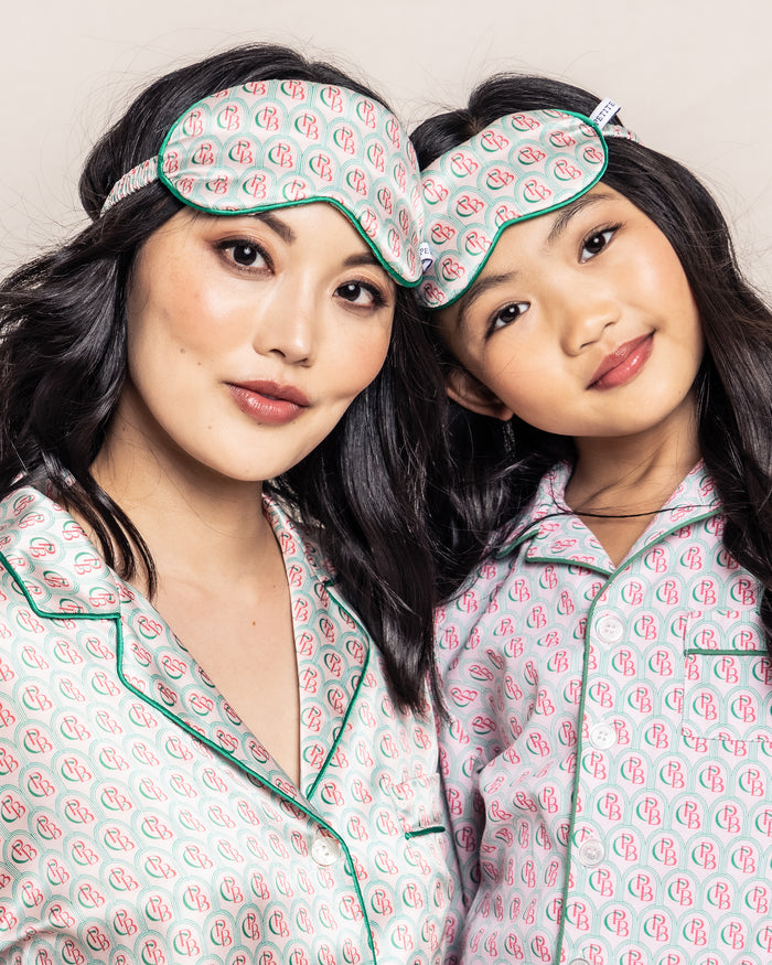 Two individuals, wearing matching Petite Plume pajamas with green piping and neutral tones, smile softly while donning the Colony Hotel x Petite Plume Kids Silk Sleep Masks in an exclusive print, capturing the serene ambiance of the Colony Hotel.