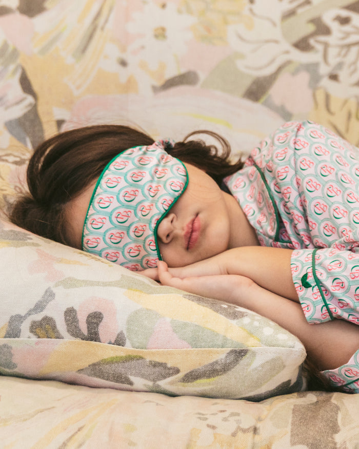 A child in Petite Plume pajamas and a Colony Hotel x Petite Plume Kids Silk Sleep Mask in Exclusive Print drifts into dreamland on a decorative pillow against a soft, patterned background.