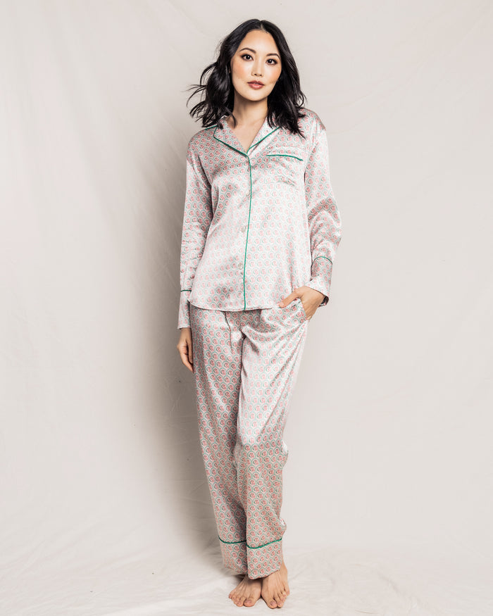 A dark-haired woman wears the Colony Hotel x Petite Plume Womens Silk Pajama Set, featuring green piping and an exclusive pattern. She stands barefoot against a plain light background, her left hand in her pocket, exuding confidence as she gazes at the camera.