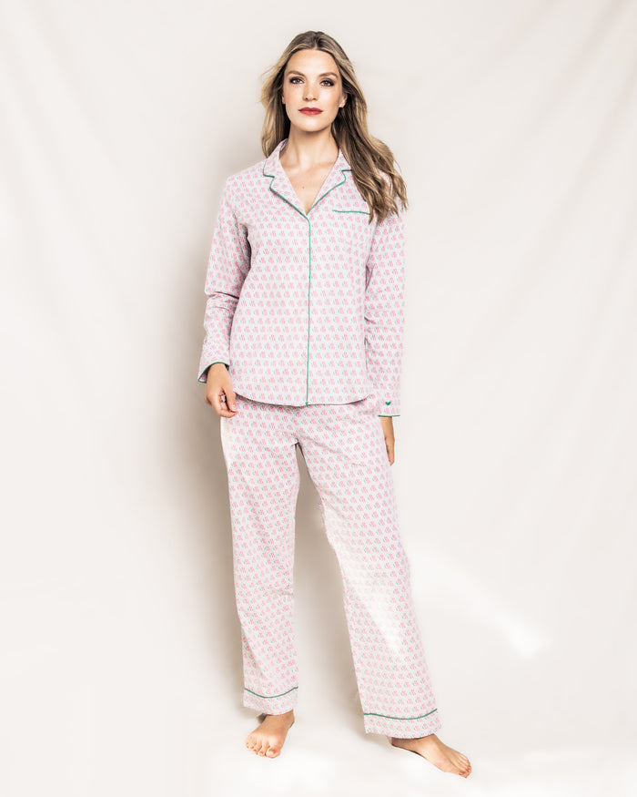 A woman with long hair stands against a neutral background, wearing a Colony Hotel x Petite Plume Womens Exclusive Print Pajama Set, featuring a button-up top and long pants. Barefoot and with one knee slightly bent, she embodies luxury sleepwear as she gazes at the camera.