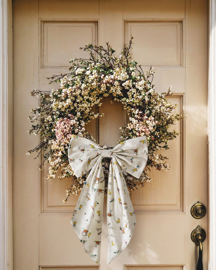 An Easter Basket and Wreath Bow in Easter Frolic with a large white bow featuring a pastel floral pattern is tied at the bottom of a wreath made of small white and pink flowers, hanging on a beige door.