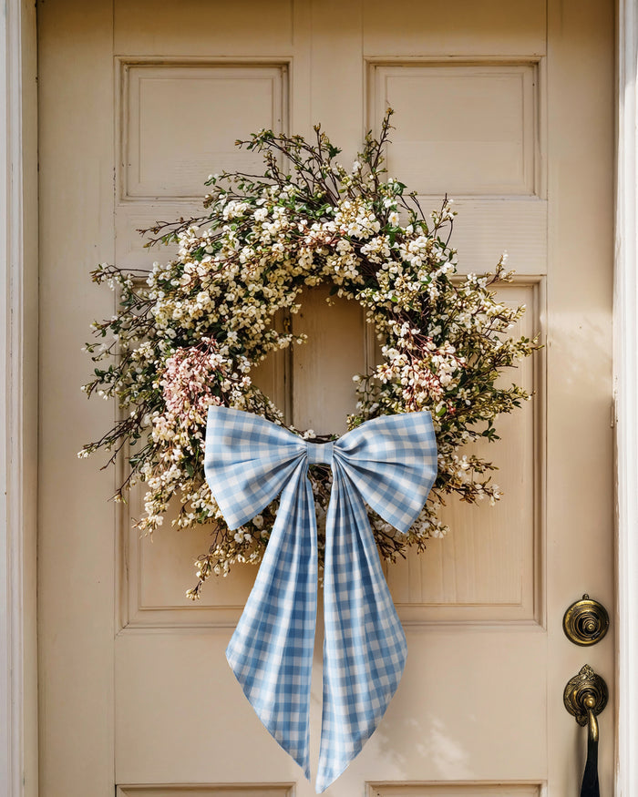 A floral wreath of small white and pink blooms hangs on a beige door, adorned with the Easter Basket and Wreath Bow in Light Blue Gingham featuring long ribbon tails.