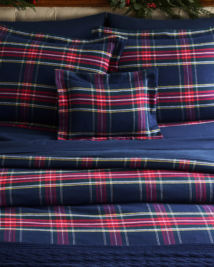 A bed featuring Petite Plumes Windsor Tartan bedding in brushed cotton flannel displays classic blue, red, and white plaid prints. Neatly arranged pillows and comforter enhance intersecting stripes, while greenery with red berries adds charm to the headboard area.
