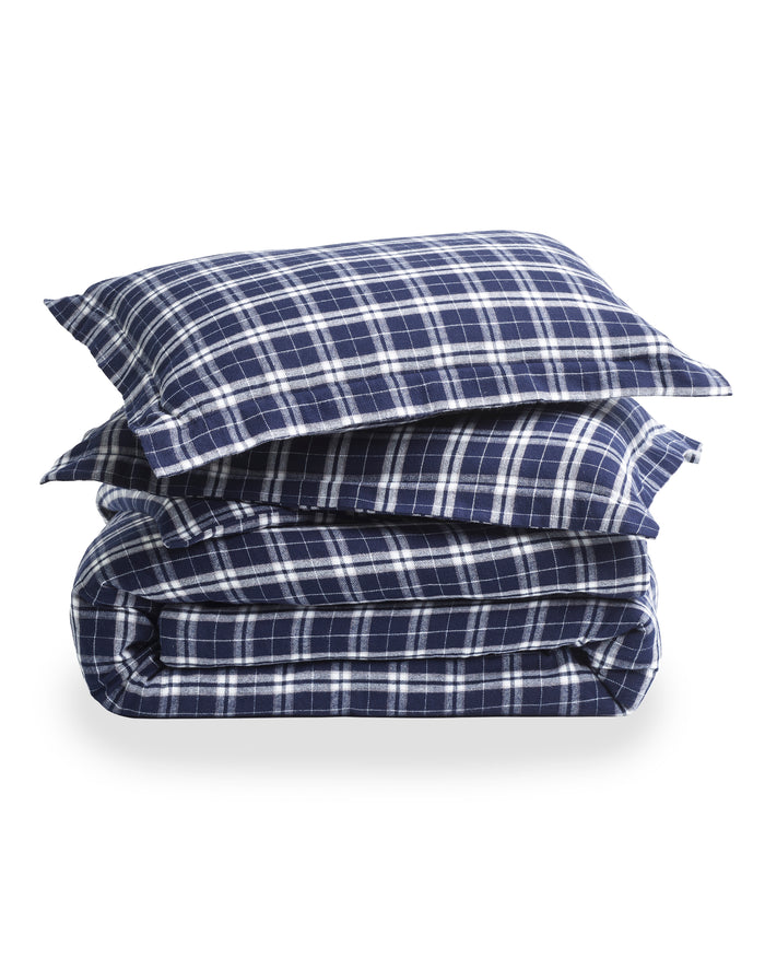 The Oxford Tartan Duvet and Navy French Ticking Bedding Set features navy blue and white plaid pillows and comforter, with striped white and gray bedsheets, next to a wooden nightstand topped with a lamp.