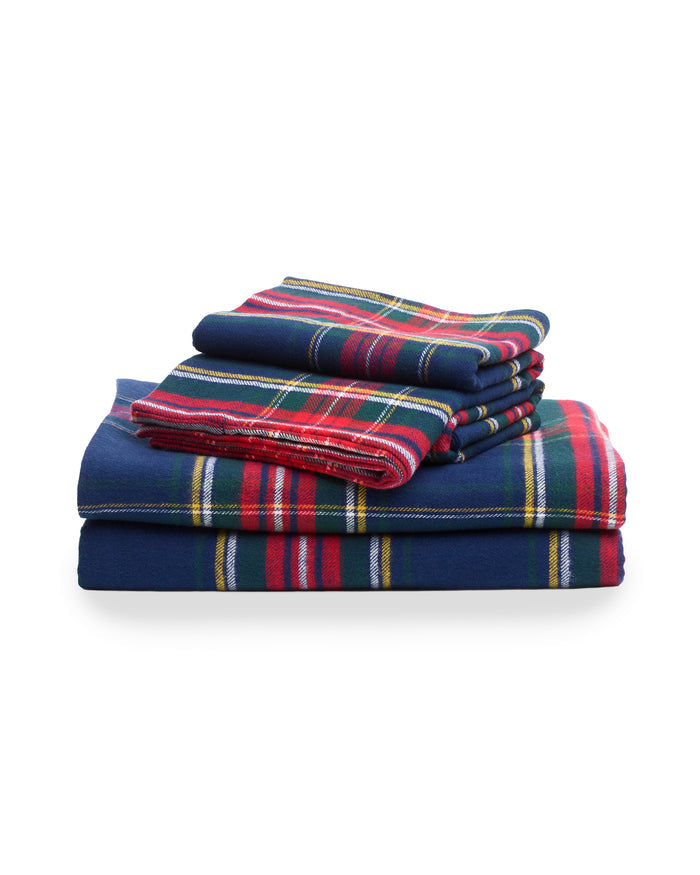 A neatly made bed showcases Petite Plumes Flannel Sheets in Windsor Tartan, featuring navy blue and red plaid cotton brushed flannel bedding with matching pillows. Set against a wooden headboard, the scene hints at a cozy, classic bedroom aesthetic.