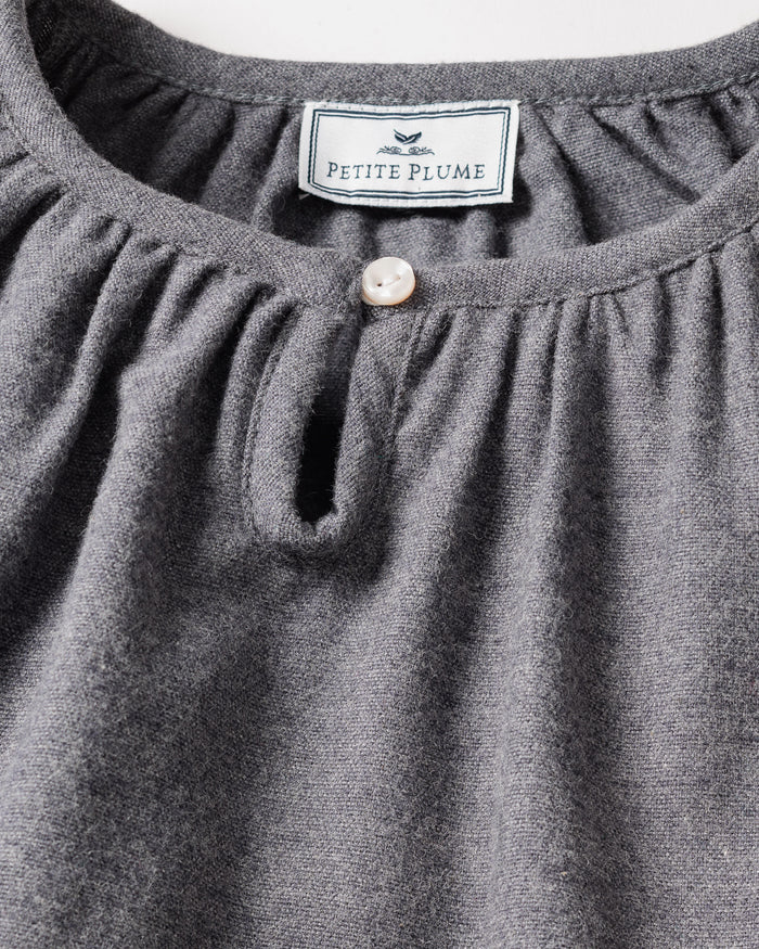 Close-up of a grey nightgown showcasing a round neckline and pearl-like button. Inside, the label reads Petite Plume with a bird graphic. Made from brushed cotton flannel, its texture highlights the softness and chemical-free comfort of the Girls Flannel Delphine Nightgown in Grey by Petite Plume.