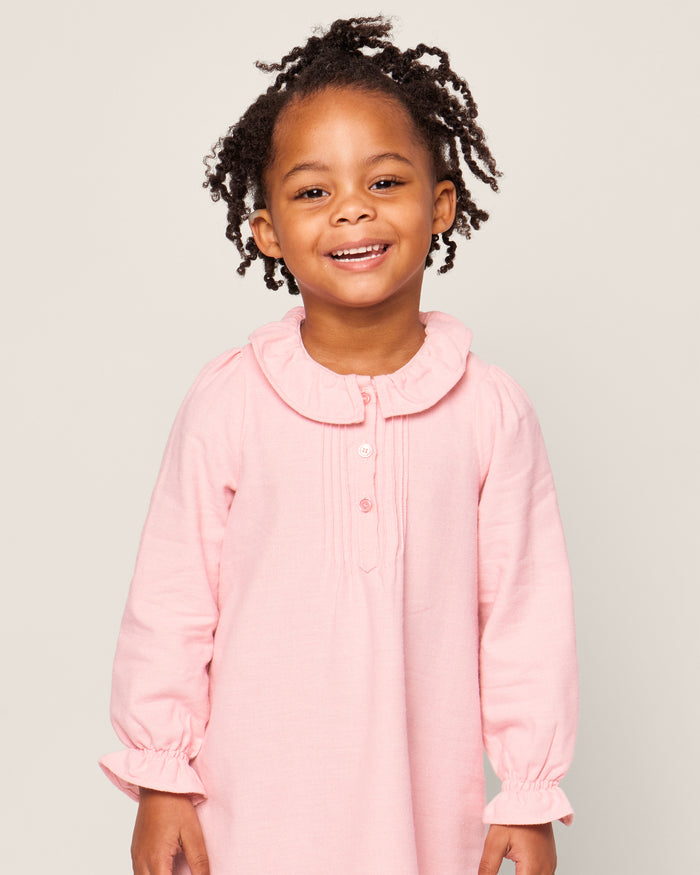 A young child with curly hair smiles in a Petite Plume Girls Flannel Victoria Nightgown in Pink. The nightgown, with a ruffled collar and front buttons, exudes charm and comfort against a plain light-colored backdrop, highlighting this elegant classic sleepwear.