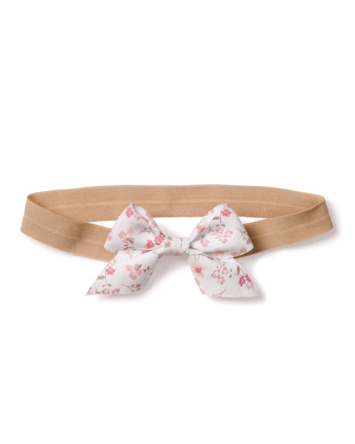 The Girls Dorset Floral Hair Bow by Petite Plume, a beige elastic headband with a white background and pink floral bow, is perfect for girls holiday gift collections as a charming stocking stuffer.