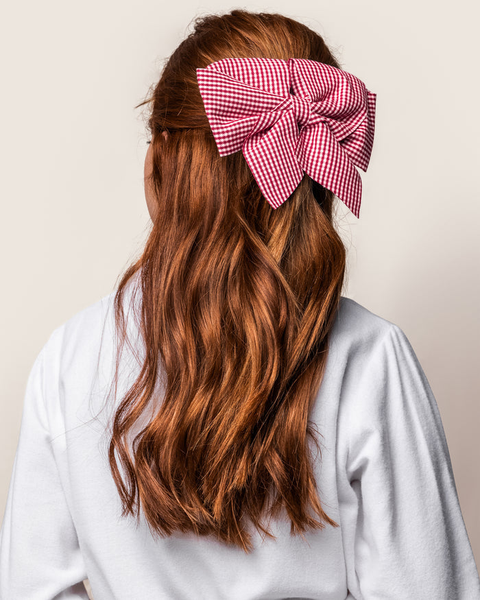 With long, wavy auburn hair, a person sports a large red and white Petite Plume Girls Hair Bow in Red Mini Gingham as an accessory. They wear a white top and face away from the camera against a light background.