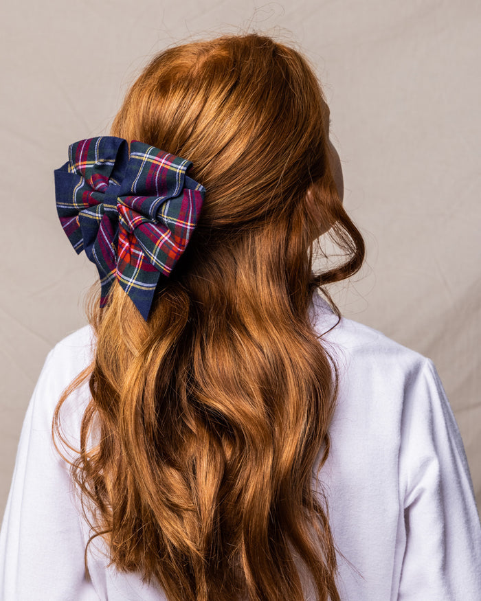A person with long, wavy red hair, tied with a large Petite Plume Windsor Tartan Girls Hair Bow in navy, red, and white, stands against a neutral backdrop wearing a crisp white shirt—ideal for gifting during the holidays.